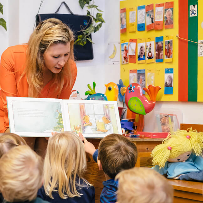 Juf Ellen leest voor uit een prentenboek. De kleuters luisteren en wijzen de prenten aan.